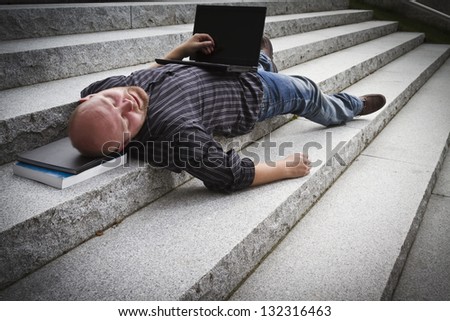 A sleeping / resting / exhausted man working with his computer / laptop in stairs outside.