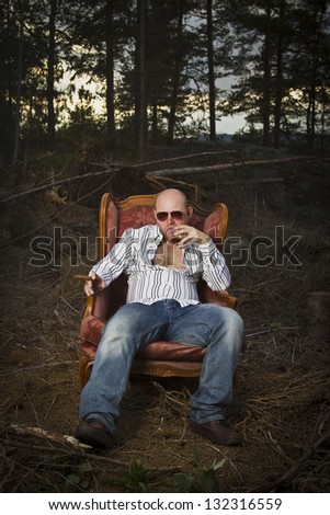 Sleazy man in a classic vintage chair in the middle of a logging area. Smoking cigar and drinking Cognac in woods.
