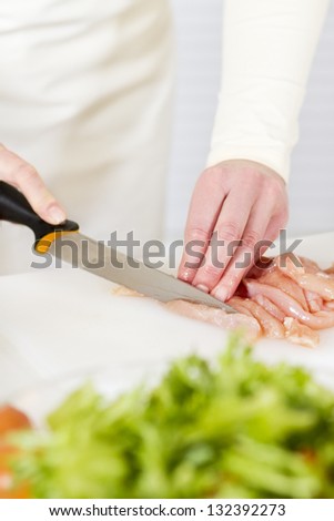 Young woman in a white kitchen chopping chicken meat fillet. Vegetable on the bench.