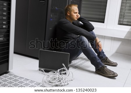 Frustrated and exhausted it engineer / consultant in a data center. Sitting apathetic in front of a server rack.