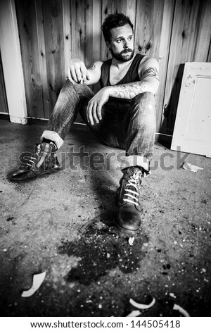 Punk rocker or redneck sits on the floor and smoke in a messy house.