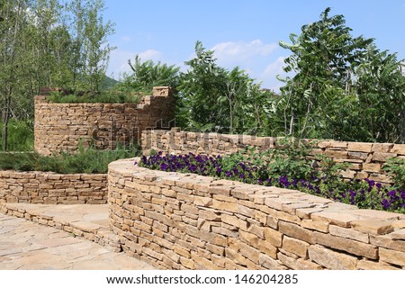 A Summer view of an ornamental garden with a slate pathway and garden wall made out of stone.
