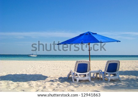 Two beach chairs under a blue umbrella on the white sandy beach at a resort.