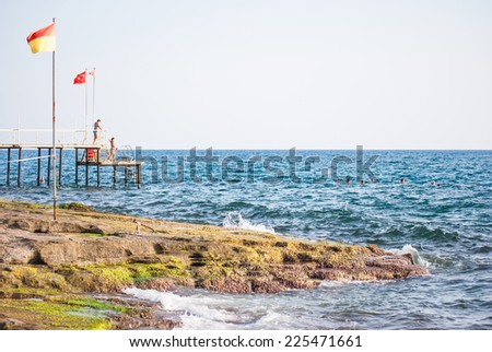 pier on the rocky sea shore, people swimming and jumping in water, turkish flag in background