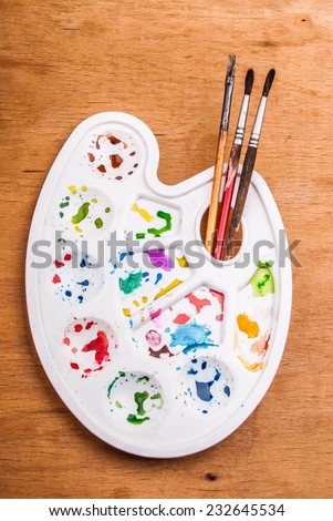 White plastic art palette with random watercolor paint blobs and brushes on wood