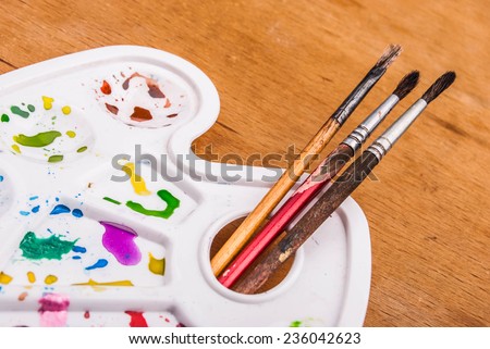 White plastic art palette with random watercolor paint blobs and brushes on wood