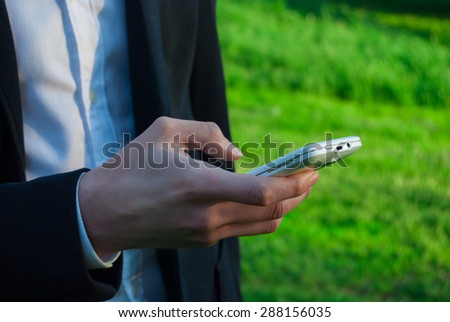 Young man wearing suit with smartphone, outdoors on the green background of trees and bushes. Business man in a green zone or park. An image for topics of finance, business and communication