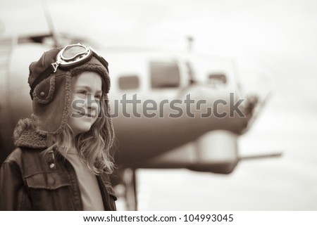 A confident young female pilot is gazing into the distance, wearing flight jacket / hat / goggles.  Bomber is visible in the background out of focus.