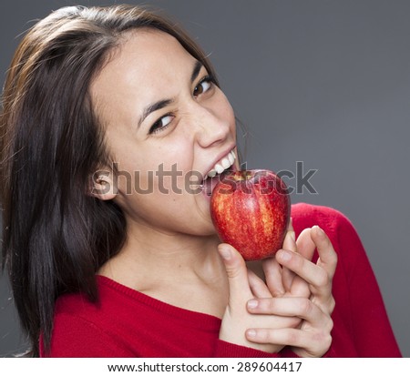happy cute young brunette with red sweater biting red apple with good appetite