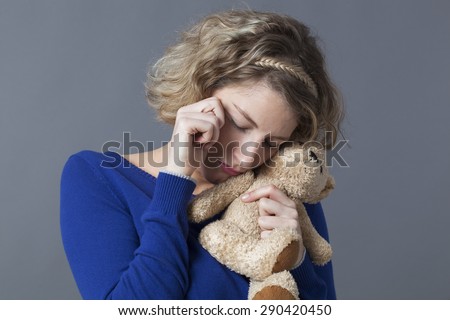 sad young blonde woman hugging her teddy bear with tenderness for emotional reinsurance and child memories
