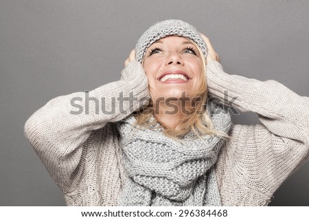 lovely young woman with blonde hair wearing winter hat and clothes, looking up with hands on ears for fun and happiness