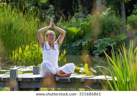 outdoors meditation - relaxed young yoga woman in praying lotus pose,closing eyes to relax and meditate on a wooden bridge with green foreground and water background, summer daylight