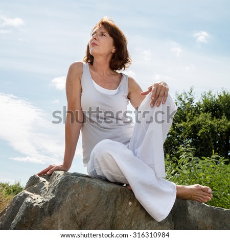 senior zen - thinking 50s woman sitting on a stone for outdoors yoga session wearing white seeking serenity and wellbeing in a park,summer daylight
