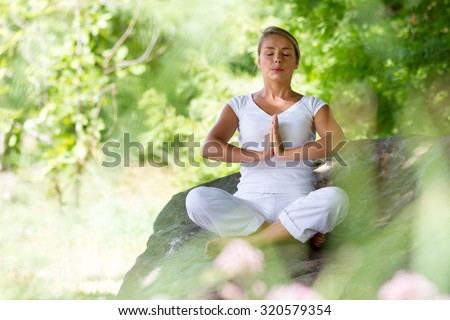 outdoors meditation - young yoga woman exercising in lotus praying pose,closing eyes to focus on inner peace to relax and meditate on a big stone,green blurred foreground and background