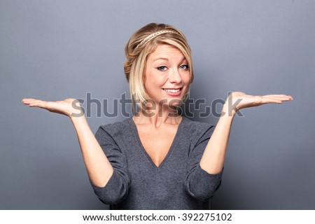 comparison concept - happy young blond woman displaying something on both flat hands for similar choice of product, gray background studio
