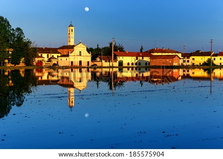 Typical Italian Landscape of a rice field with Houses and Rice Paddy  with water. Blue Sky with moon and the houses, mirrored on the Rice Paddy lightened by the sunset with bright yellow color.