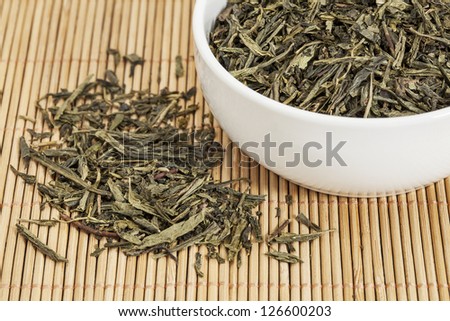 loose leaf Sencha green tea in a white china cup and spilled over bamboo mat