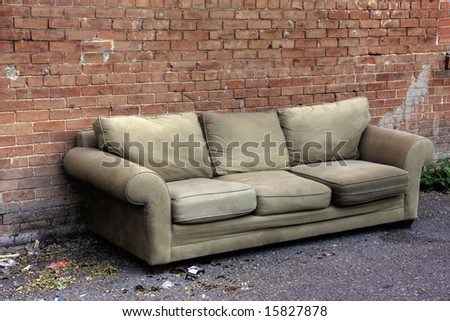old sofa discarded in an alley, surrounded by trash and broken glass, against red brick wall