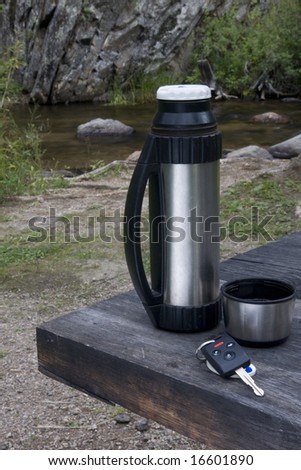 car key, steel thermos bottle and cup of tea on a roadside picnic table with scenic mountain stream in background