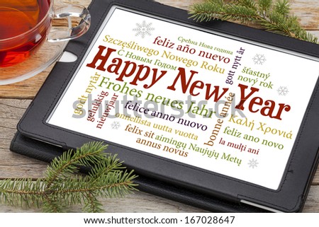 Happy New Year in different languages on a digital tablet with a cup of tea and spruce twigs