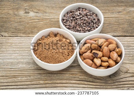 cacao beans, nibs and powder in white ceramic bowls against grained wood