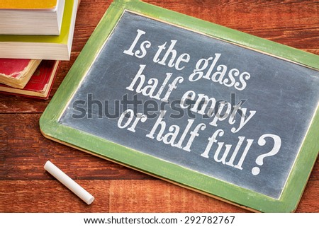 Is the glass half empty or half full question on a slate blackboard with a white chalk and a stack of books against rustic wooden table