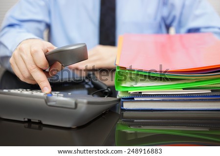 Businessman is dialing  phone for advice when he got a lot of files. Assistance is needed
