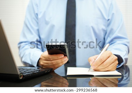 busy businessman at  office desk is using smart mobile phone, writing in notebook and watching computer screen