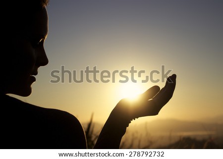 Young woman silhouette at sunrise cups rising sun in hand, landscape background