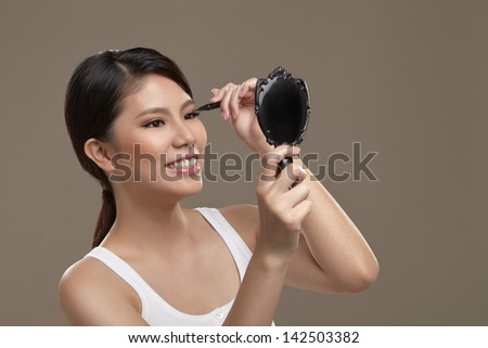 A female asian with natural make up holding a mirror and liquid eye liner applying eye liner to her eye lid while looking at a mirror