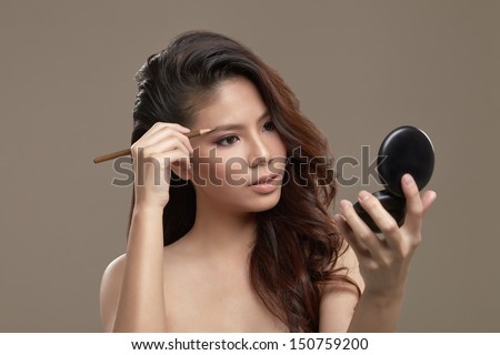 A female asian with make up and pink eye shadow holding an eye brow pencil drawing eye brow while looking at a mirror