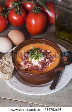 Tomato salmorejo soup in a ceramic bowl with the raw ingredients, tomatoes, eggs, oil and bread over wooden background