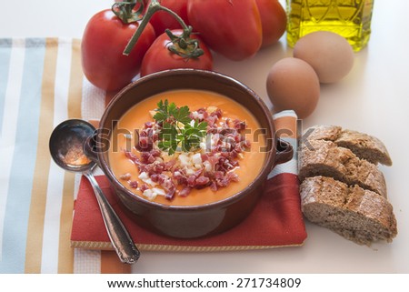 Tomato salmorejo soup in a ceramic bowl with the raw ingredients, tomatoes, eggs, oil and bread over white background