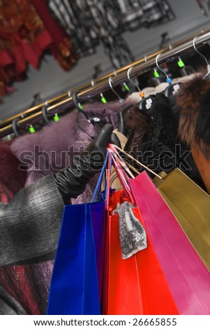 Hand holding a bunch of shopping bags in front of a clothes stall in a shop. A piece of cloth hangs out of one of the shopping bags.