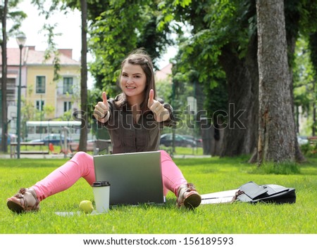 Happy young woman with a laptop rising her thumbs up, outside in an urban park.
