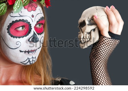 Portrait of young blond girl in black dress with Calavera Mexicana makeup mask and with skull in her hand. Halloween