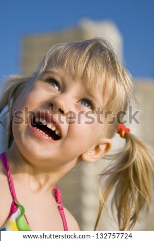 Cheerful blonde girl with two tails enthusiastically looking at the sky at the beach on a sunny day, close-up,  against the blue sky and tall buildings, cities