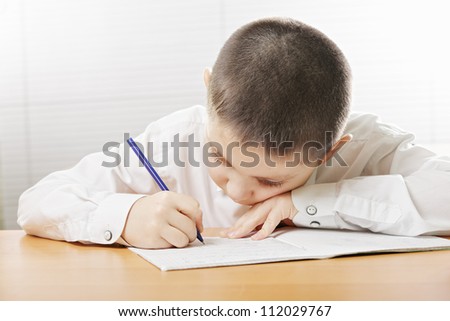 Boy in white shirt writing at desk bending over copybook
