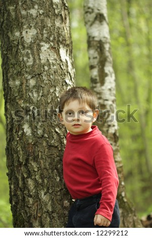 Boy in red jacket standing at tree at forest looking straight