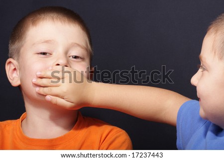 Boy shuts brother's mouth by palm over dark background