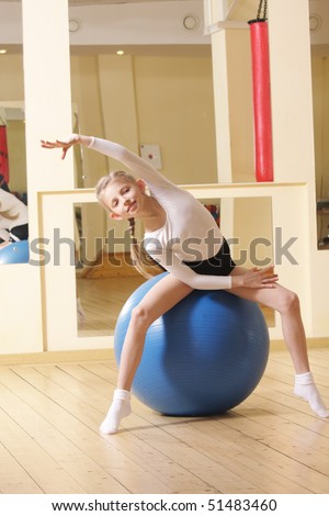 Little gymnast girl sitting on big blue ball in gym bending right side