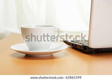 White cup in saucer and laptop on desk closeup photo