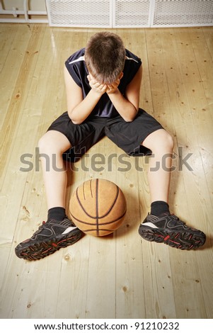 Kid with basketball sitting on hardwood floor covering face with hands