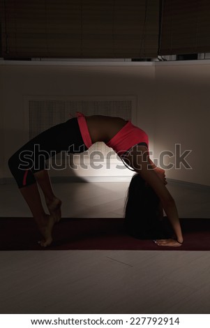 Young brunette woman exercising back bend yoga pose in dark gym