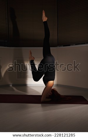 Redhead woman exercising head stand pose one leg strighted and another bended in dark gym