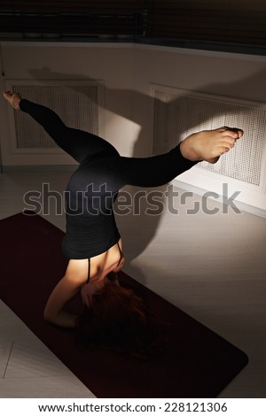 Redhead woman exercising head stand with split pose in dark gym