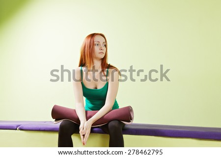 Caucasian redhead woman sitting at gym with rolled mat on knees