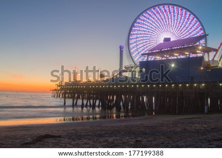 Sunset on the Santa Monica Pier in California. Angled view includes ocean, sand, Santa Monica pier, lit ferris wheel, and orange sunset.