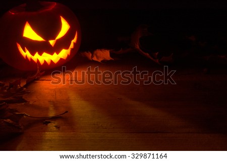 Spooky halloween background with face of jack o lantern in the corner with red shadows on the wooden surface with autumn dried leaves