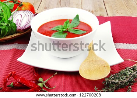 cold fresh diet tomato soup with basil thyme and dry pepper in big bowl over red mat on wood table ready to eat